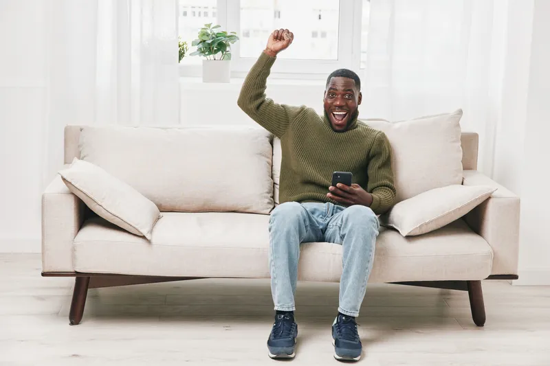 Excited black man celebrating success on smartphone while sitting on a modern couch in a bright, cozy living room