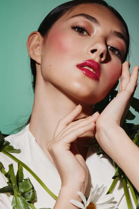 Young woman with glossy red lips and shimmery eyeshadow poses elegantly against a green background. Fashion and beauty concept with fresh floral accents.