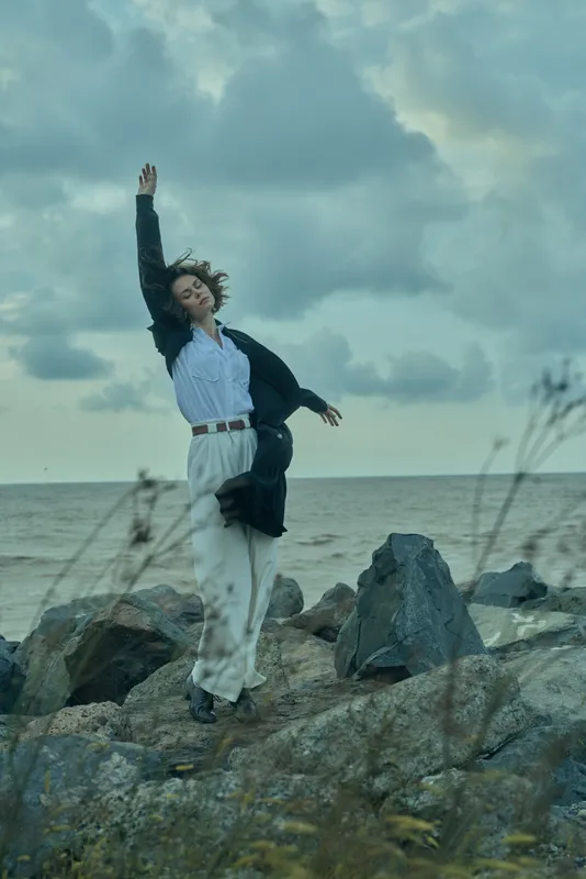 Joyful leap on a rocky shore with the sea in the distance, wind tousling hair, a smart blazer and light trousers expressing freedom and style in an outdoor coastal scene