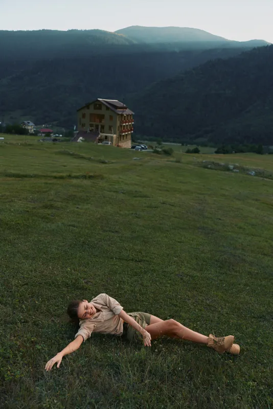 A woman lies on a grassy field outdoors, distant house and mountains in the background, conveying relaxation, leisure, and a moment of nature inspired calm.
