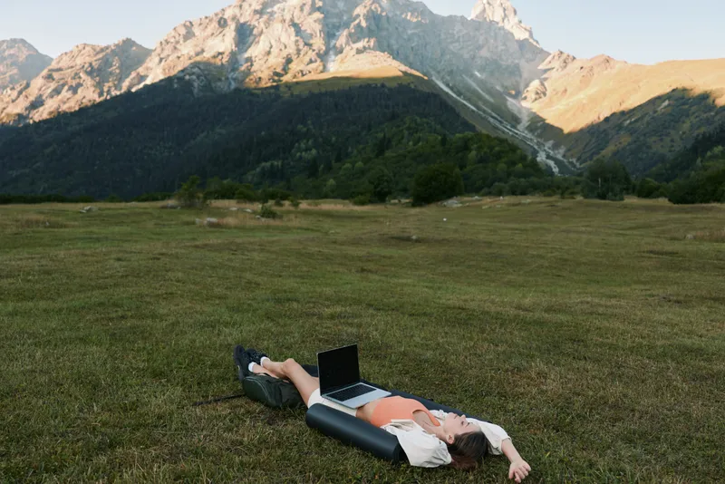 A person lies on a grassy meadow with a laptop, set against distant mountains under a clear sky, creating a calm remote work moment in nature.