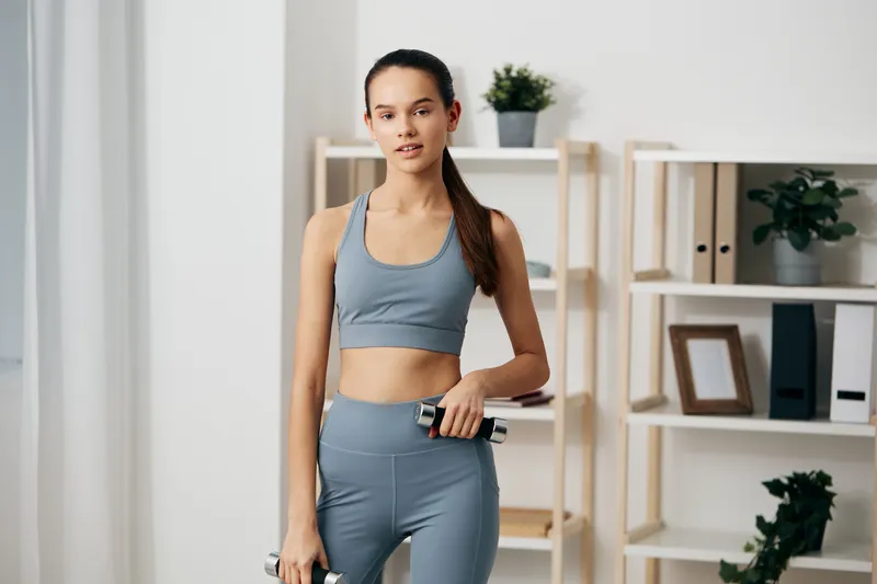 Woman fitness, sport, home gym, workout, dumbbell, athletic, space, fitness equipment, training, exercise: A woman in blue athletic wear stands in a bright home gym with a dumbbell in hand, ready for