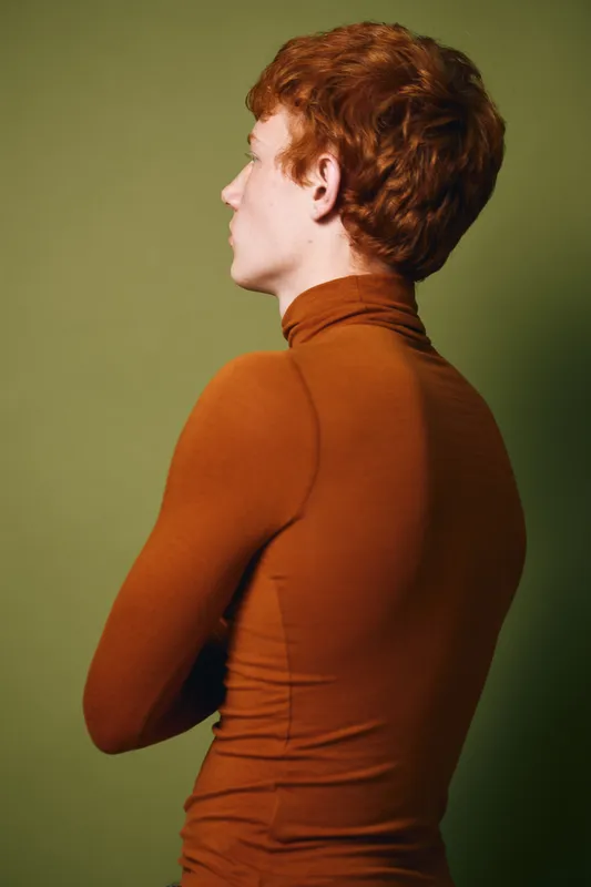 Young man with red hair wearing brown turtleneck sweater standing with arms crossed and looking sideways on green background in studio portrait fashion style.