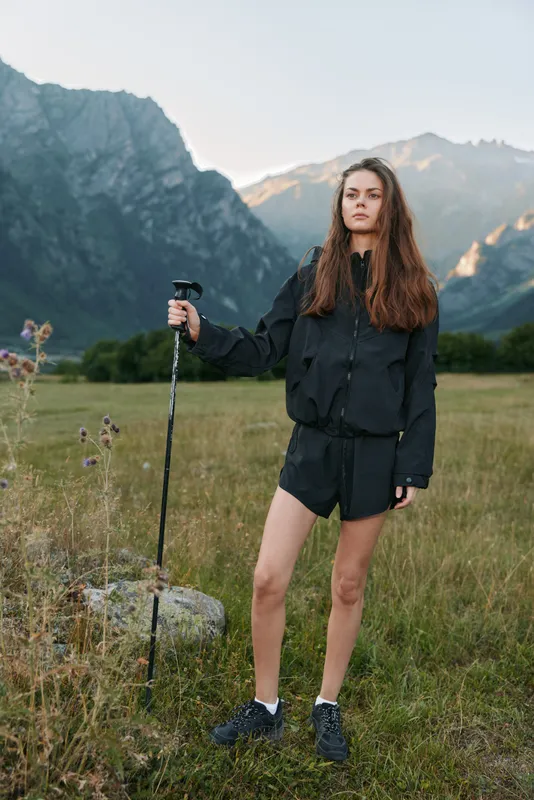 A young woman stands in an open field with a trekking pole, framed by rugged mountains. She wears a black jacket and shorts, ready for outdoor adventure.