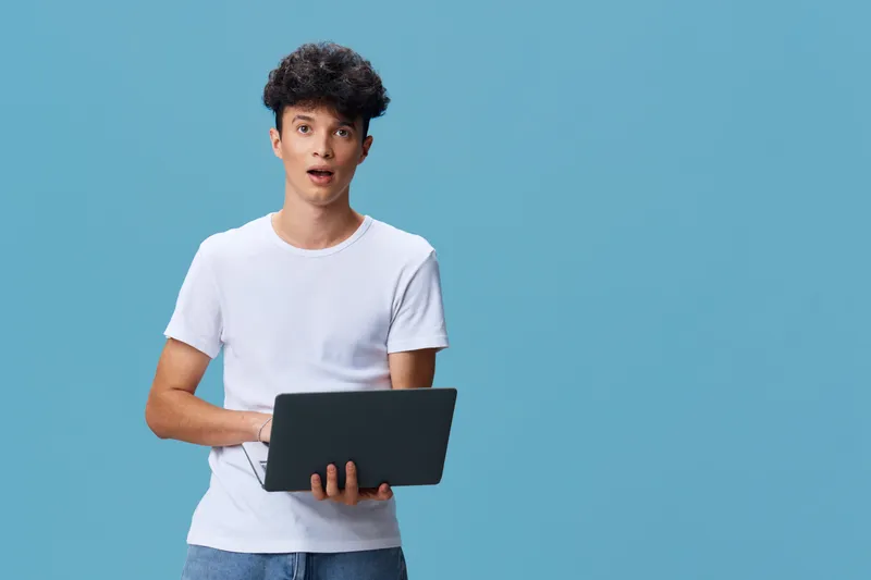 surprised young man holding laptop wearing casual white t-shirt and jeans with curly hair standing against blue background in modern tech concept.