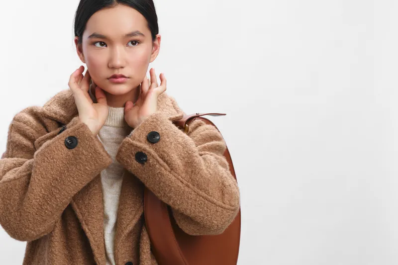 asian woman adjusting her coat in a clean studio setting, conveying calm confidence and poised style, as soft light highlights her thoughtful expression.