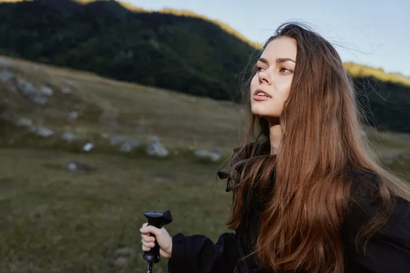 A young woman stands outdoors in a rugged landscape, gripping a trekking pole and gazing into the distance with calm confidence, capturing a mood of adventure.