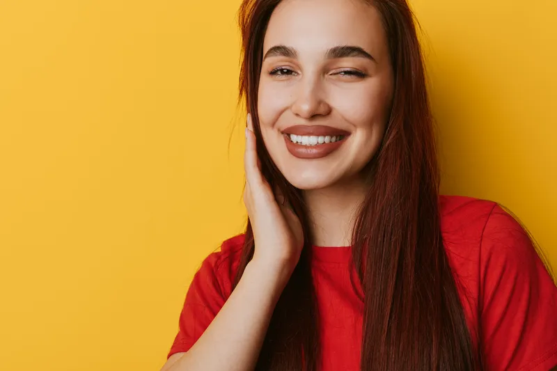 Young woman with long hair smiling and touching face in red shirt against yellow background. Portrait of happy female with natural makeup and confident expression in studio lighting.