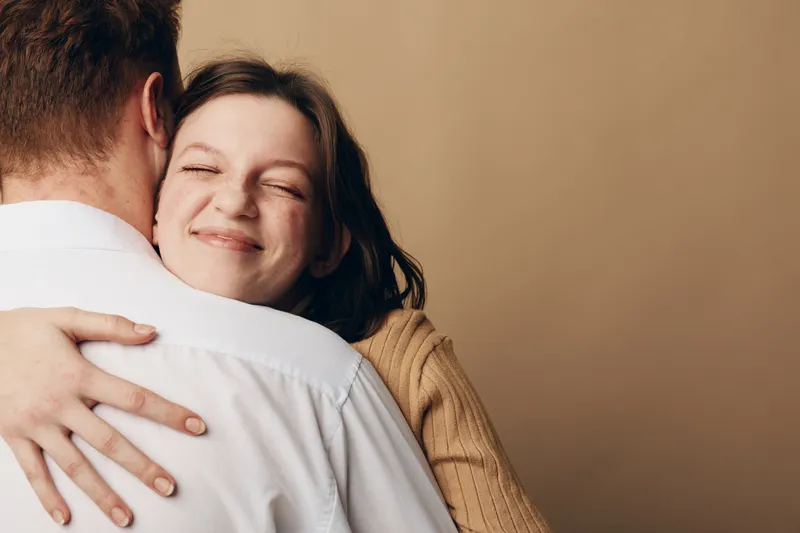 Happy young woman with closed eyes hugs man in white shirt, expressing affection and warmth. Soft brown background creates cozy and intimate atmosphere of love and connection.