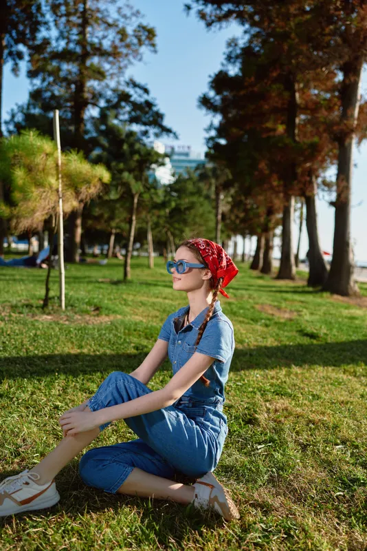 Lifestyle woman in modern boho-western denim street style sits on grass wearing sunglasses and a red bandana with natural trees and cityscape background in film color.