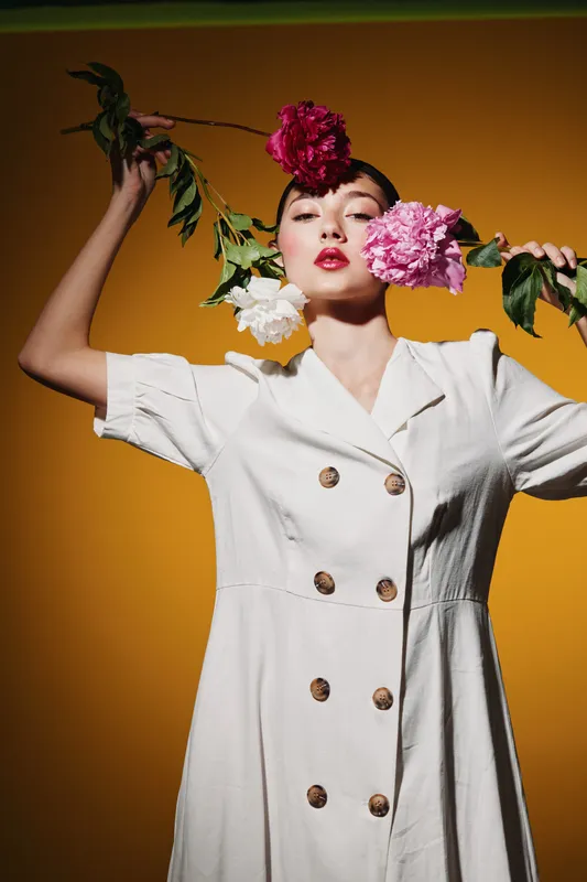 Stylish young woman in a white buttoned dress holds vibrant pink and white flowers, posing creatively on a warm yellow background. Concept of fashion, beauty, and floral creativity.