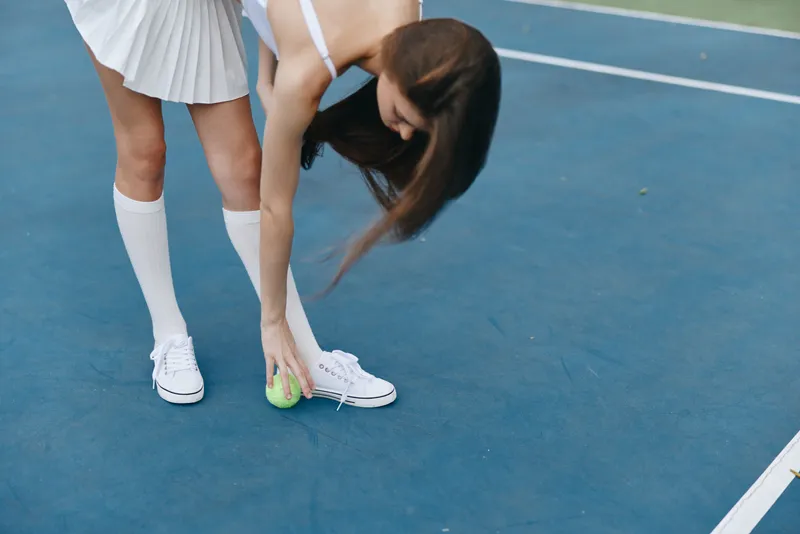 Young woman in a tennis outfit bends to pick up a green ball on a blue court, exuding focus and determination, reflecting the sport s competitive spirit, showcasing athleticism