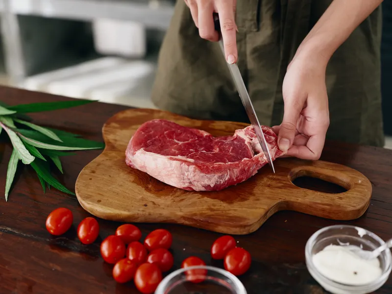 Женщина с ножом в руке нарезает свежий стейк из мяса для жарки на кухне