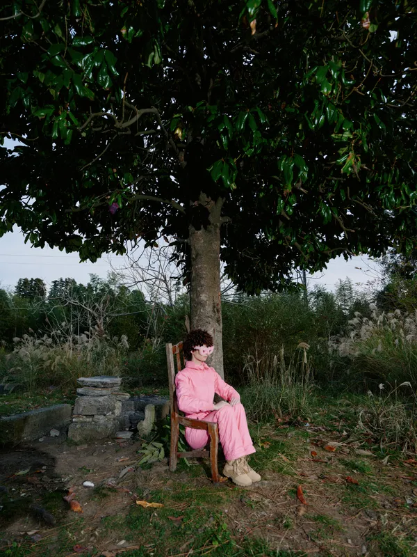 A person sits on a wooden chair beneath a large tree in a rural setting, wearing a pink outfit and beige shoes, enjoying shade and calm as nature surrounds a quiet moment outside.