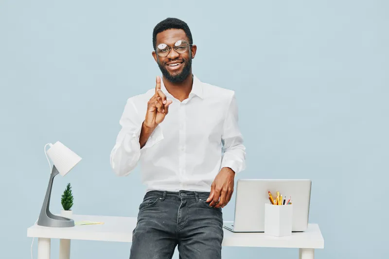 Happy confident man with beard and glasses standing in modern office, smiling and gesturing with index finger, wear white shirt, relaxed pose, workspace with laptop and supplies, professional business