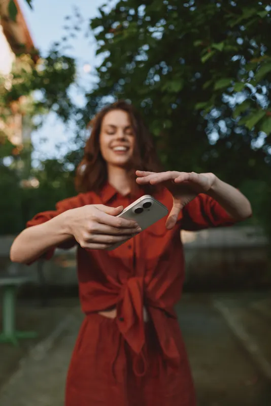 Smiling woman in a red dress enjoying a casual outdoor moment, taking a selfie in a garden setting. Lively lifestyle concept with warm natural light and cheerful mood