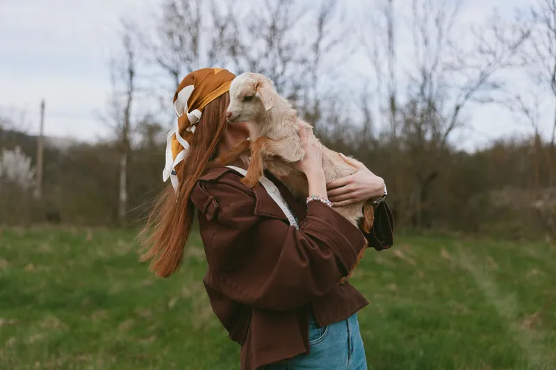 Young woman outdoors holding a goat kid close, brown jacket and scarf, field background. Lifestyle moment focused on animal care, joy, and nature in a countryside setting. Soft daylight enhances