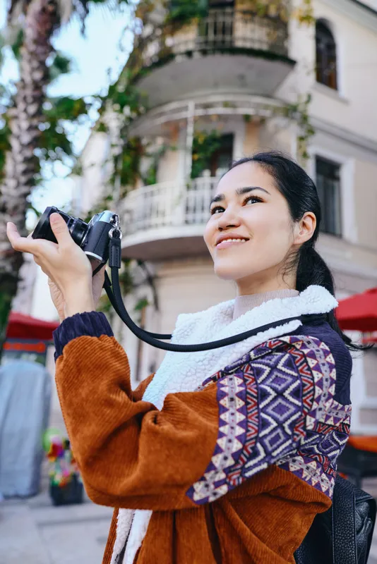 Camera in hand woman captures urban street scenes outdoors showcasing photography travel and city life with a warm smile colorful jacket and scarf during a casual shoot