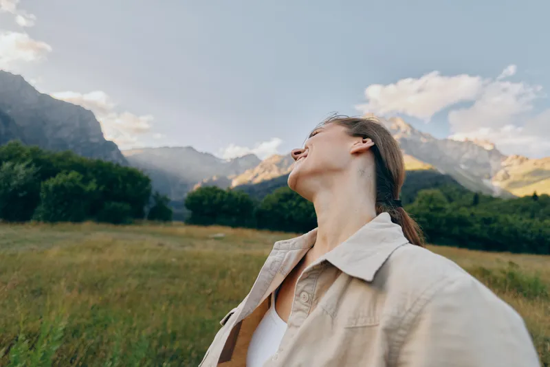 Young woman enjoys peaceful moment in nature with mountains and blue sky in background wearing casual beige jacket and knitted hat in outdoor lifestyle scene.