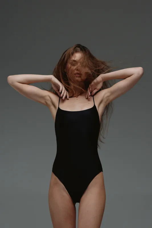 slender woman in black swimsuit posing with hair covering face against gray background