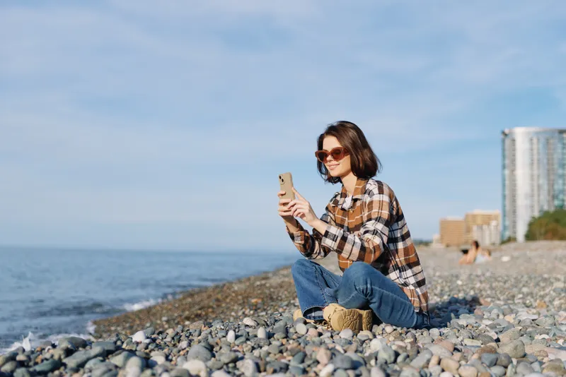 Casual woman sitting on a pebbled beach with a smartphone and sunglasses, relaxed by the sea, wearing a plaid shirt and jeans, city skyline in the distance, natural light and coastal leisure.