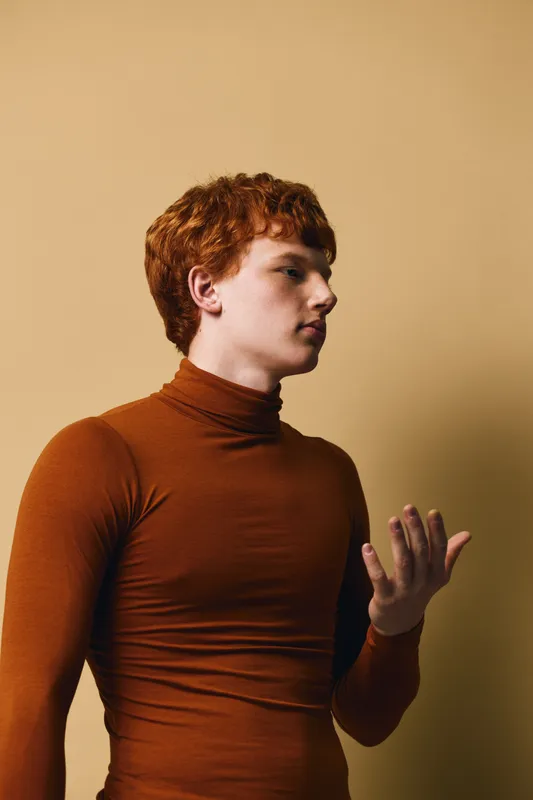 Portrait of a young man with red hair wearing a brown turtleneck sweater against a plain beige background. Side view, thoughtful and casual expression.