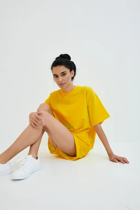 Young woman in bright yellow casual outfit sitting on white floor, looking relaxed and calm in minimalistic bright studio setting.