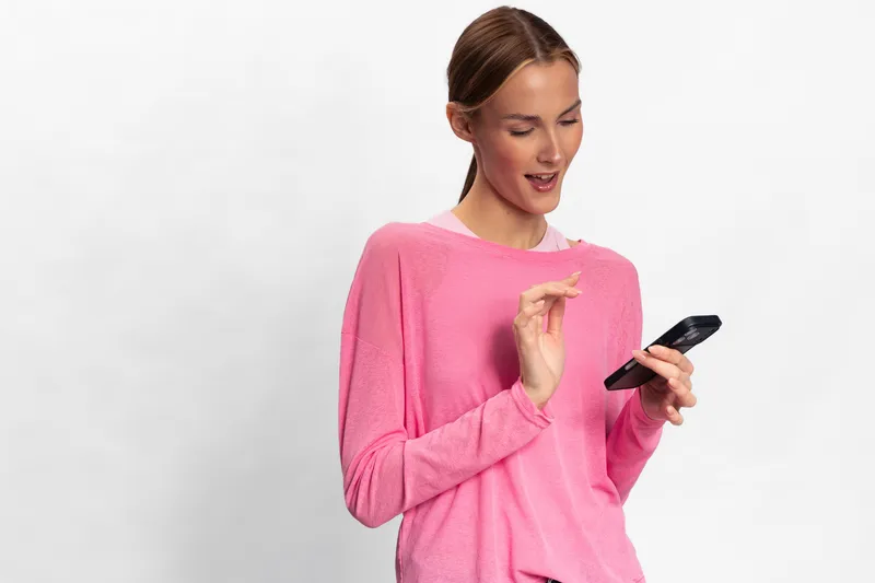 Young woman in pink sweater uses smartphone with smile. She stands against white background with focused expression. Concept for technology, communication, social media, and lifestyle.