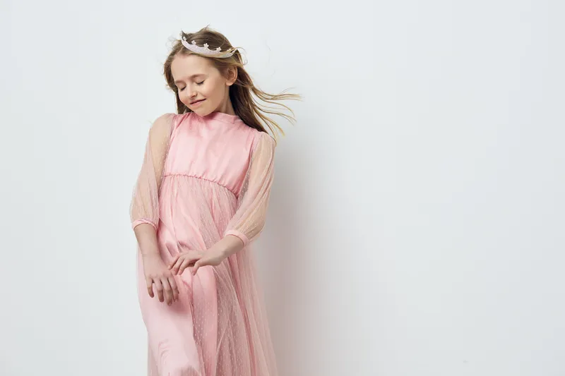 Young girl in a pink dress with flowing sleeves, smiling gently in a bright, minimalistic background, embodying joy and innocence in childhood fashion
