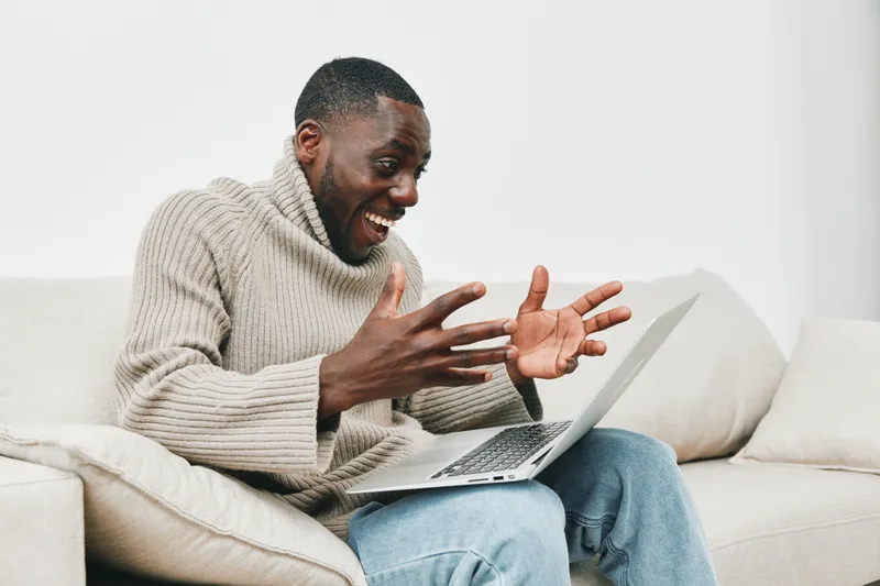 Happy African man wearing a beige sweater, celebrating success while using a laptop in a bright room, conveying excitement and joy with expressive gestures
