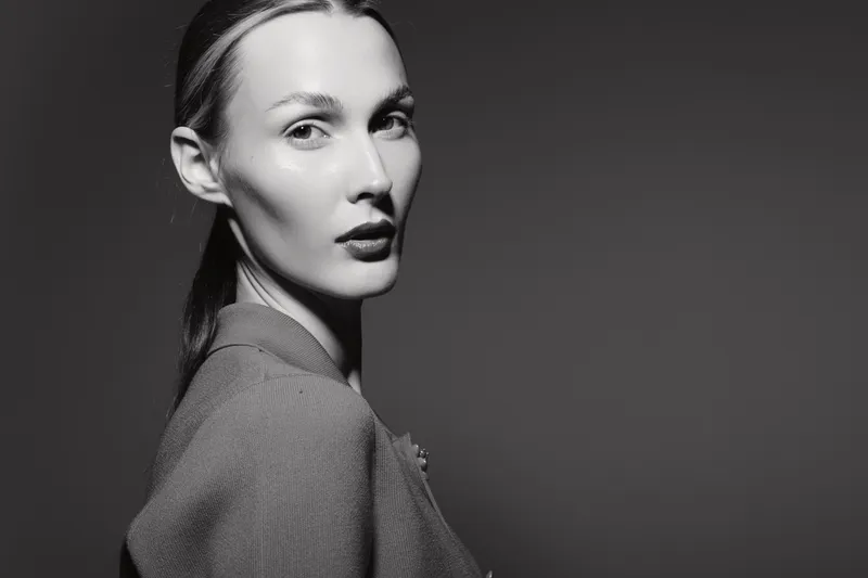 Portrait of a young white woman with light skin and slicked back hair wearing textured clothing. The black and white image highlights facial features and fashion in studio lighting.