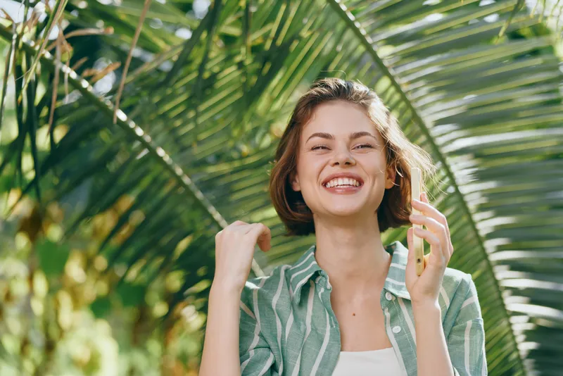 Woman smiles brightly outdoors in a lush tropical setting. A confident, relaxed pose conveys joy and vitality, ideal for lifestyle, travel, and wellness narratives with sunshine and greenery.
