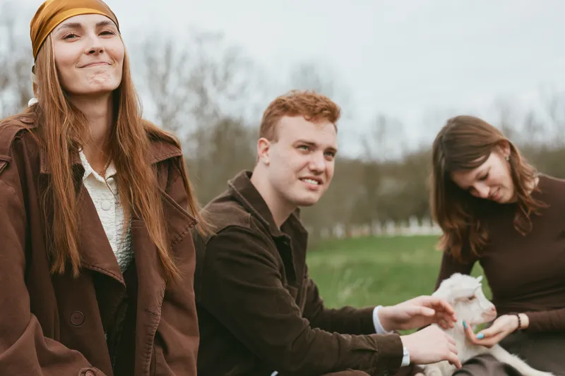 Relaxed countryside lifestyle scene with friends outdoors and a playful kid goat, smiling faces, casual jackets, green field backdrop, warm interaction, calm mood, simple everyday moment in nature.