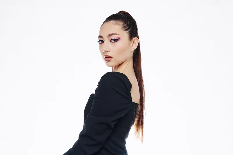 Young woman with long ponytail wearing elegant black dress poses in studio against white background with creative pink eye makeup. Concept of beauty fashion and modern style.