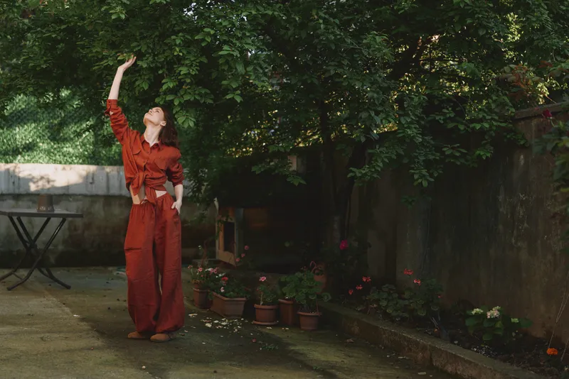 Young woman in a red jumpsuit stands in a sunlit garden reaching toward the leafy branches with a relaxed smile, capturing a casual outdoor lifestyle moment