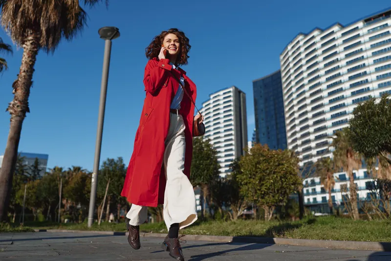 Young woman with curly hair walks confidently on a city sidewalk wearing a long red coat and white pants while smiling and talking on her phone on a sunny day.
