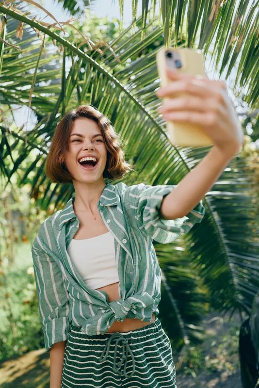 A joyous woman takes a sunny selfie among palm fronds, wearing a green striped shirt, capturing a vibrant, summery moment in tropical light.