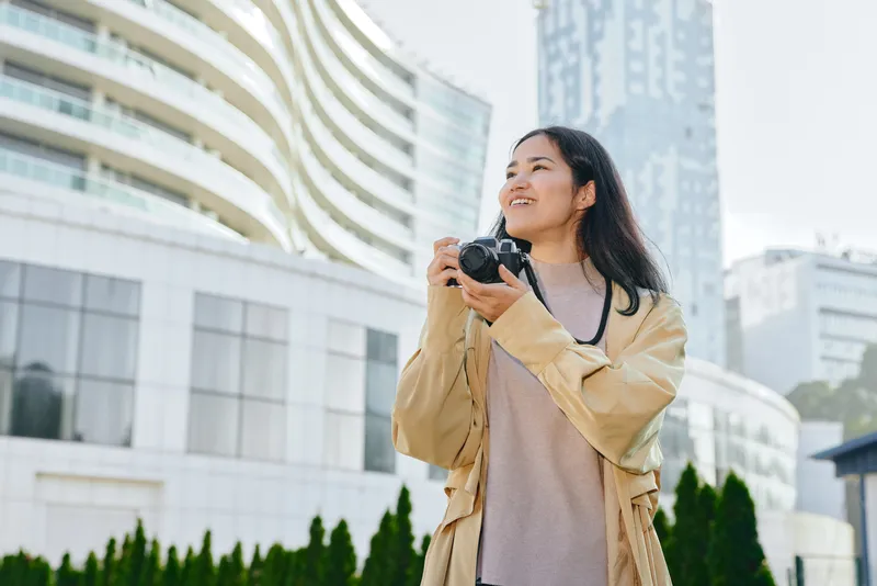 Woman holding a camera outdoors in a modern city, casual coat and sunny weather, urban backdrop with glass towers and greenery, photography in progress