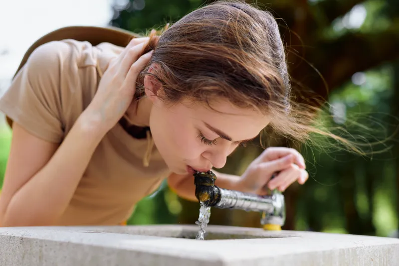 Woman girl drinking water from outdoor fountain in park, leaning over concrete basin, summer casual clothing, natural daylight and green background, refreshment and hydration captured in candid