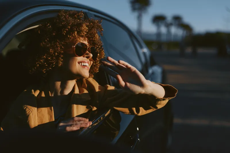 Smiling woman with curly hair leaning out of a car window. Warm sunlight and retro film tones convey lifestyle, travel, and happiness in a candid moment.