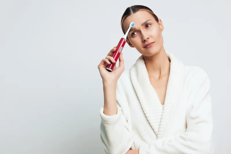 Young woman in white bathrobe holding electric toothbrush near temple. Concept of dental hygiene, morning routine, personal care, and healthy lifestyle. Soft lighting on neutral background.