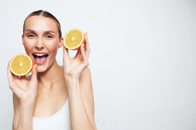 Bright young woman with a cheerful smile holds two orange halves near her face, standing against a plain light background for a fresh, healthy moment captured in a bright minimalist setup.