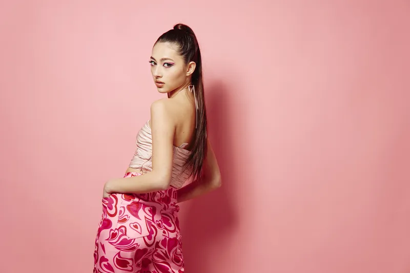 Fashionable young woman with long ponytail poses confidently against pink background wearing vibrant creative outfit. Concept for beauty, creative fashion, style inspiration.