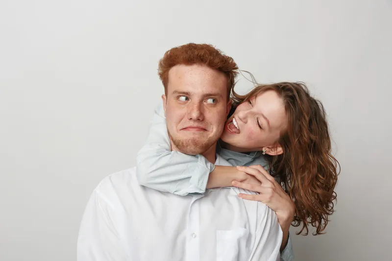 Playful young woman hugs surprised man from behind, both with casual clothes on neutral background. Concept of friendship, fun, affection, and joyful human connection.