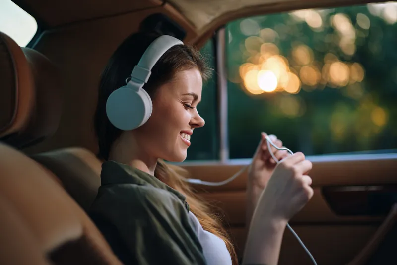 Young woman enjoying music in car with headphones, smiling, nature background, golden hour light, lifestyle concept, relaxation, happiness.