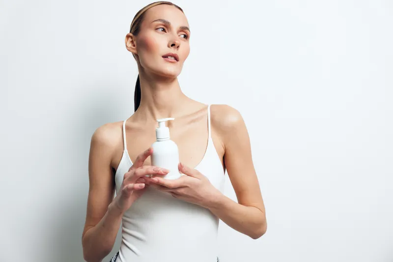 A young woman applying lotion with a pump bottle in a bright studio, showcasing a calm skincare routine and confident daily self-care.