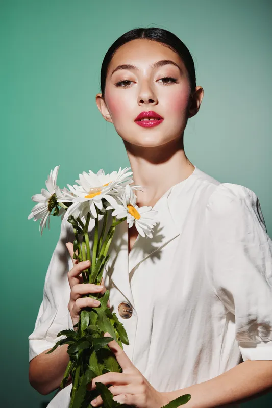 Young woman with glossy skin and bold red lips holds white daisies in a minimalistic studio. She wears a simple white top, representing beauty, fashion, and creativity.