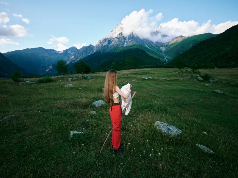 Mountains, meadow, hiker, outdoors, expansive landscape with rolling field and distant peaks, a figure in red pants holding a jacket, capturing a peaceful moment in nature