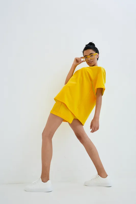 Woman in yellow casual outfit wearing matching sunglasses and white sneakers posing confidently against a plain white background in a studio setting.