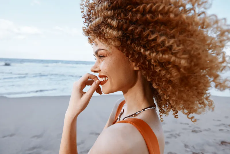 Радостное лето на берегу моря: счастливая молодая модель с кудрявыми волосами в бикини улыбается и смеётся, наслаждаясь природой и свободой у синего моря